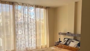 Een slaapkamer met een bed, houten vloer en een raam met dunne gordijnen.