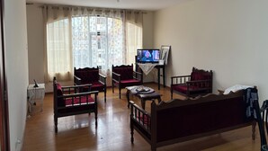 Een woonkamer met een houten vloer, twee rode fauteuils, een houten bank, een televisie op een dressoir en een raam met dunne gordijnen.