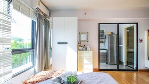 Quarto de hotel moderno com cama, bandeja com planta, cozinha visível através de uma porta de vidro e vista para o verde lá fora.