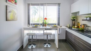 Uma cozinha moderna com bancada branca, dois banquetes, uma janela com persianas e um vaso com flores vermelhas.