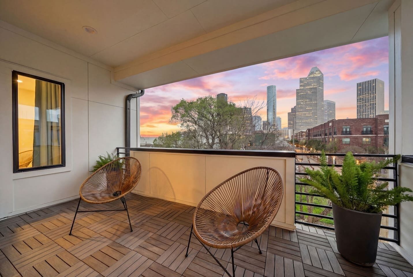 Un balcon avec deux fauteuils en osier, une plante en pot et une vue sur la ville au coucher du soleil.