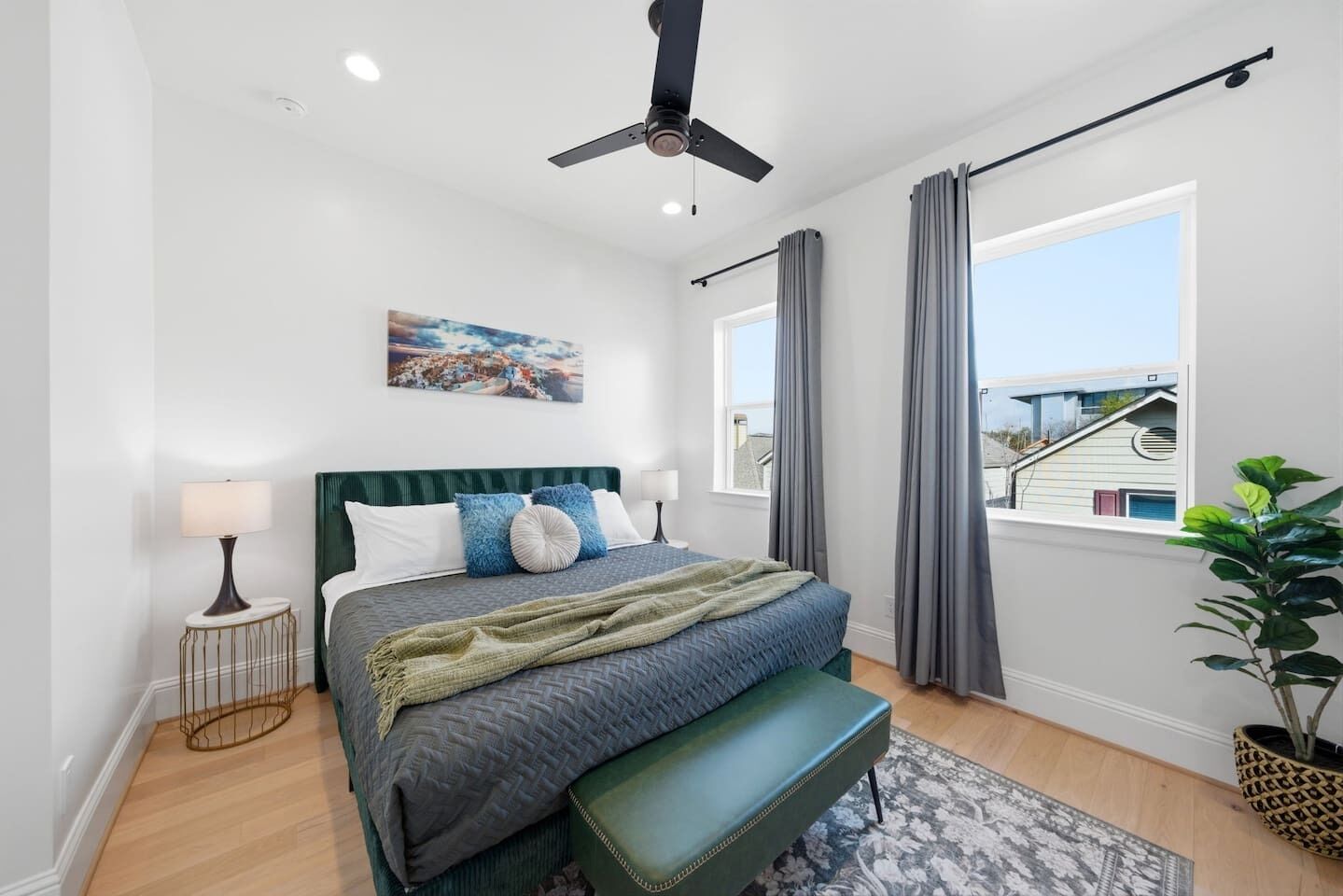 Une chambre à coucher avec un lit, un ventilateur de plafond, une fenêtre avec des rideaux, une lampe de chevet et un tableau au mur.