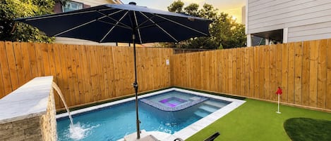 A backyard with a swimming pool, a wooden fence, a patio area with a chair, and a red flag.