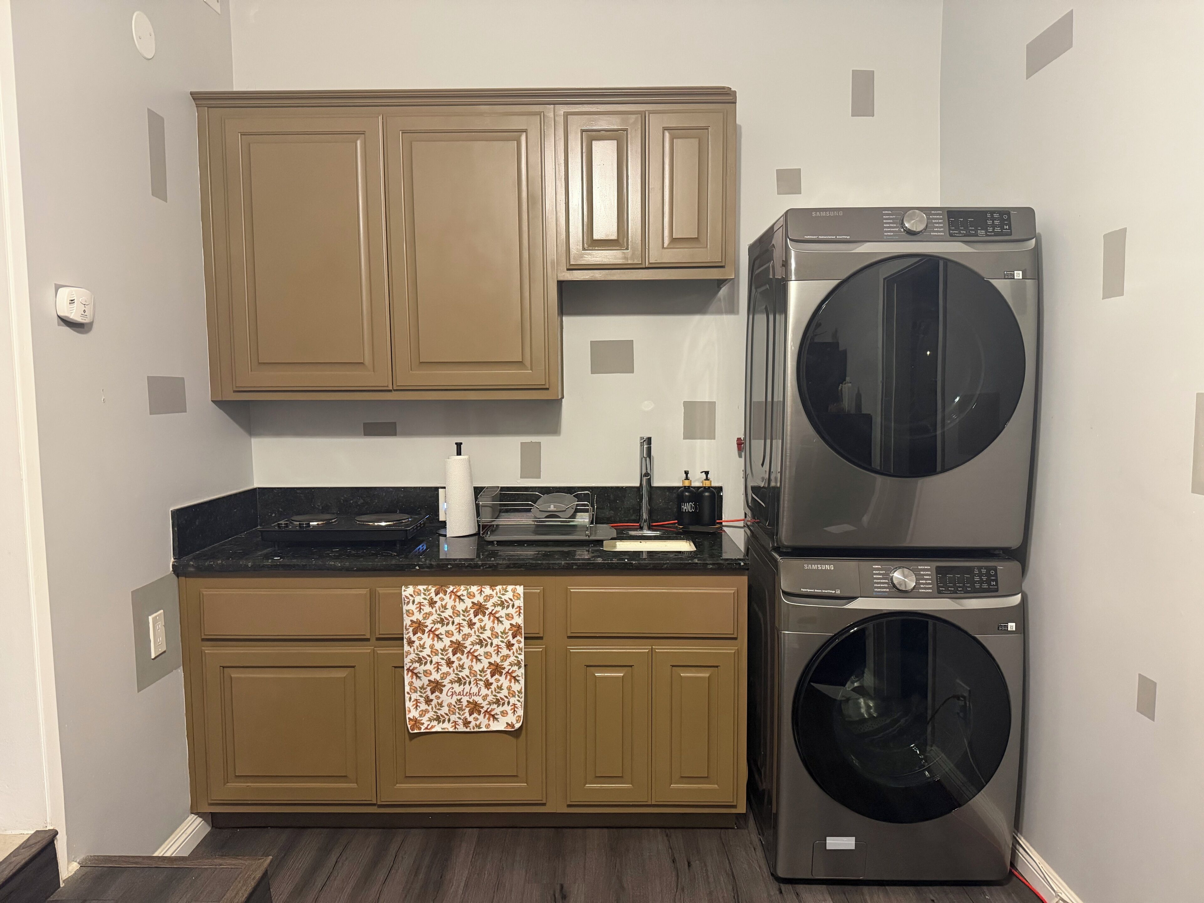 A compact kitchen with wooden cabinets, a stainless steel washer and dryer, a countertop with a paper towel holder, and a towel with a floral pattern.