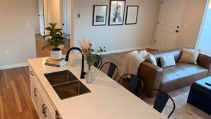 Ett modernt kök med en central köksö, en matplats med stolar, en soffa och ett soffbord. På väggen hänger inramade tavlor och på köksön står en växt.