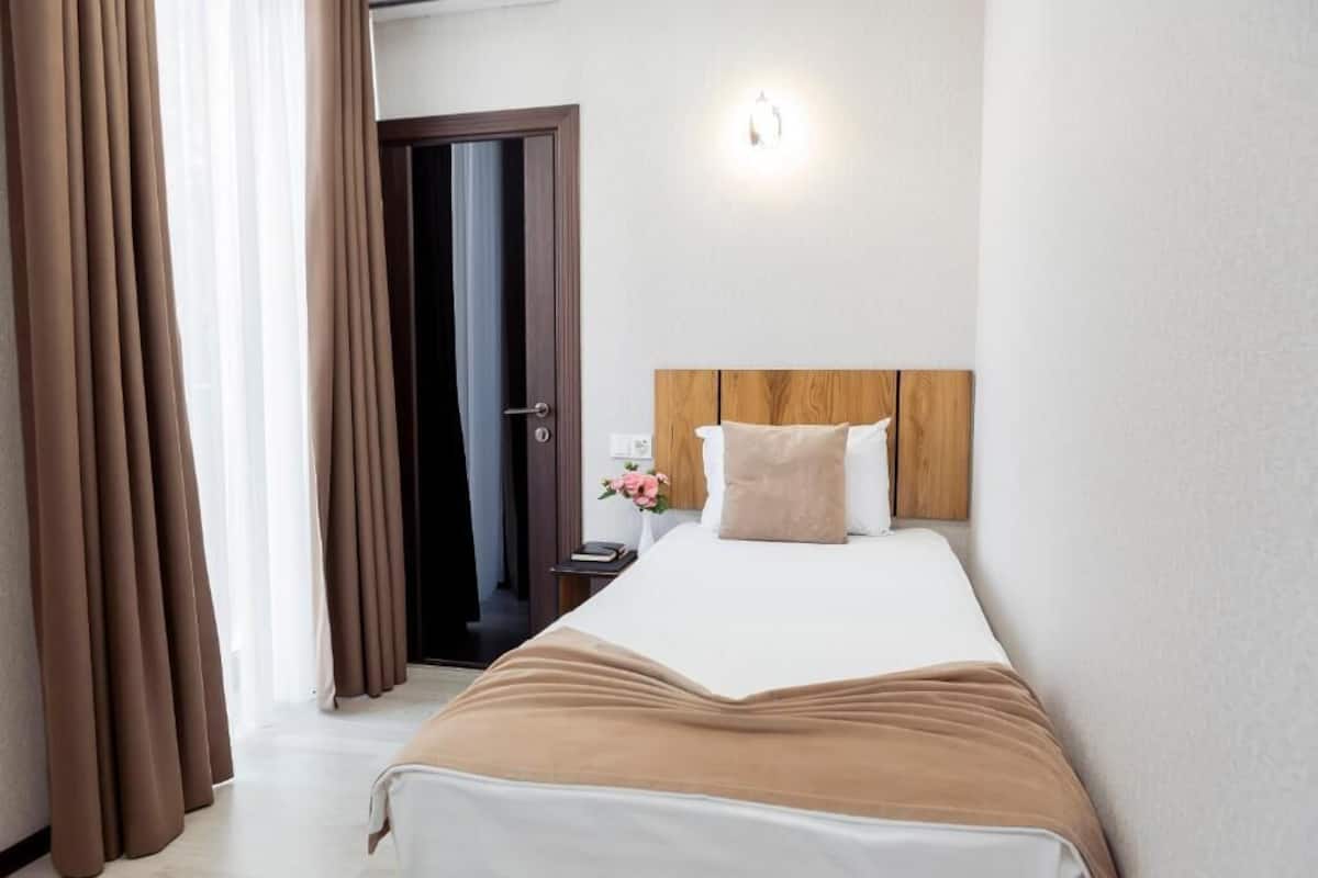 Una habitación de hotel con una cama, cabecera de madera, mesita de noche con lámpara y una puerta que da a otra habitación.