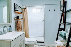 Une salle de bain avec un lavabo blanc, des toilettes, une douche avec une porte en verre et des étagères contenant des articles de toilette.
