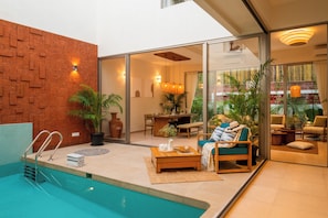 A modern indoor pool area with a clear glass wall, a brick wall, and a seating area with a table and chairs. - TBK VILLA  VAGATOR R4 (Vagator)