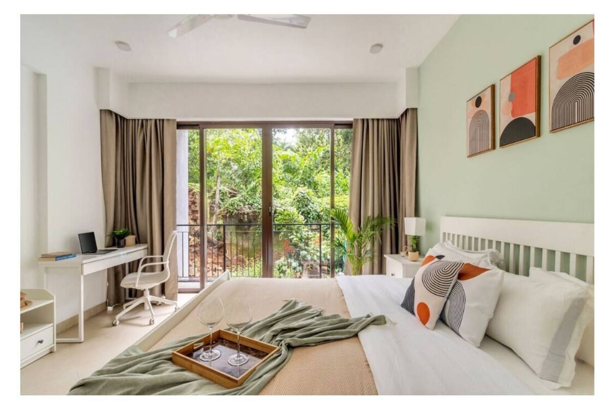 A hotel room with a large bed, a desk with a laptop, a chair, and a balcony with a view of greenery.