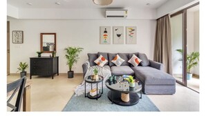 Ein modernes Wohnzimmer mit einem grauen Ecksofa, einem schwarzen Couchtisch und einem schwarzen Beistelltisch. Zwei Blumentöpfe, ein gerahmtes Bild an der Wand und ein Deckenventilator sind ebenfalls zu sehen.