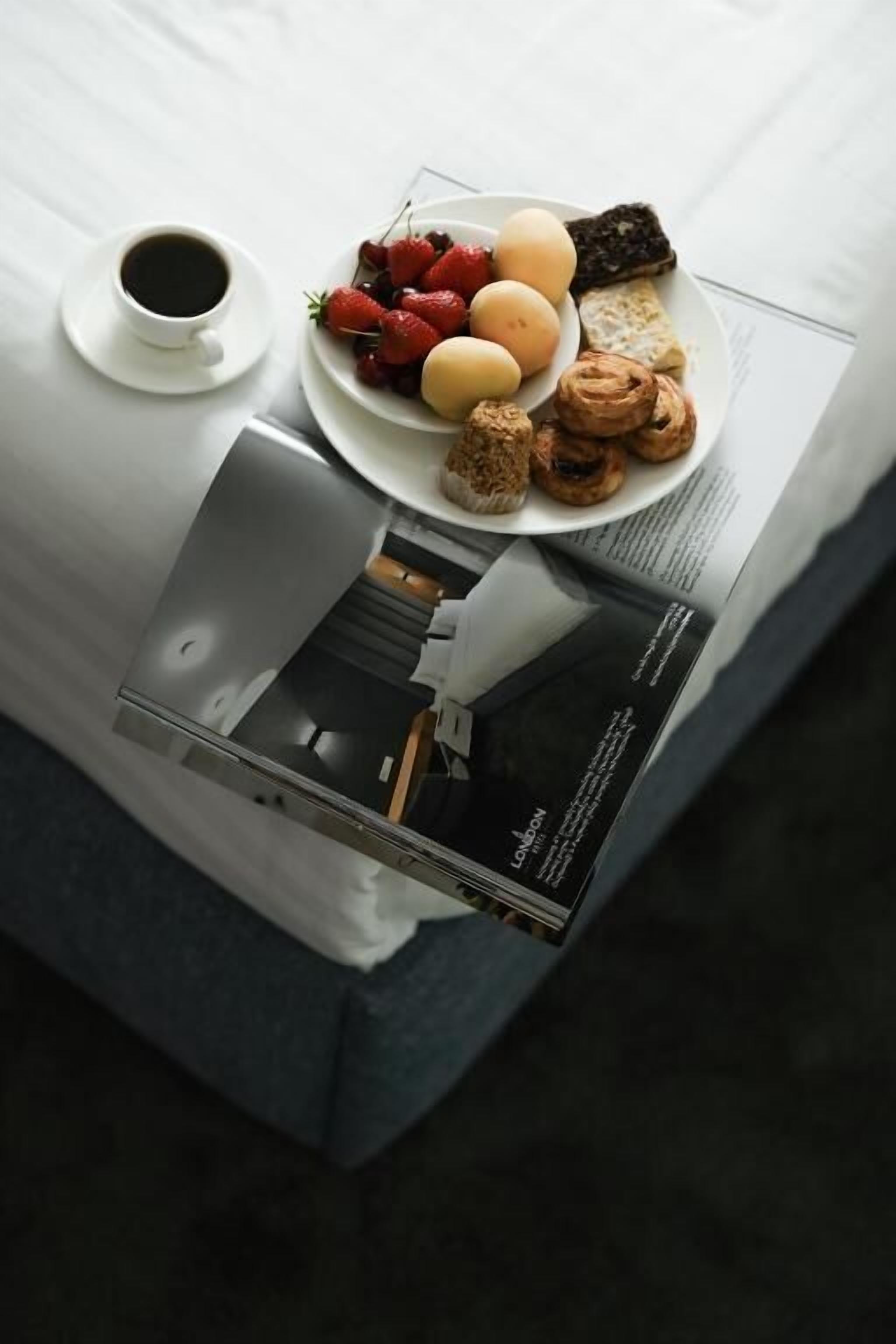 A breakfast setup with a plate of strawberries, eggs, and pastries, a cup of coffee, and a magazine on a bed.