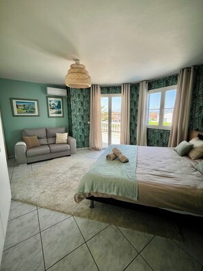A bedroom with a bed, a sofa, a hanging light fixture, and a view through a window.