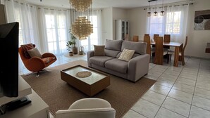 Un salon moderne comprenant un canapé, un fauteuil, une table basse, un téléviseur et un coin repas avec des chaises et un lustre.