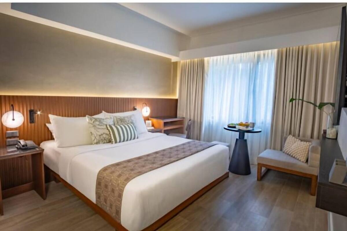 Egy modern szállodai szoba, melyben egy nagy ágy, éjjeliszekrények, egy gyümölccsel tálalt kis asztal, egy kanapé és nagy ablakokon keresztül a külső tájra nyíló kilátás található.