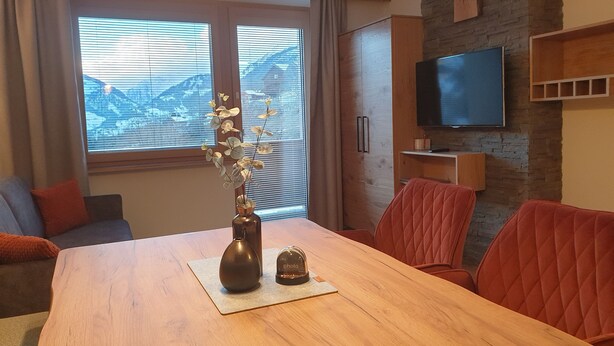 Ett modernt vardagsrum med ett matbord i trä, röda stoppade stolar, en platt-TV som är monterad på en tegelvägg och ett stort fönster med persienner.