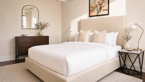 A bedroom with a large bed, a dresser, a mirror, a painting, and a nightstand with a lamp.