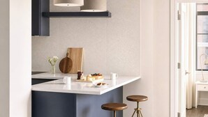 A modern kitchen with a white countertop, two bar stools, a wooden cutting board, and a hanging light fixture.