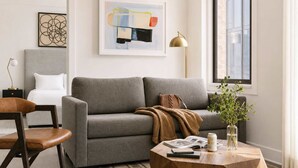 A living room with a grey sofa, a wooden coffee table with books and a plant, a side table with a lamp, and a framed abstract painting on the wall.