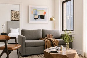 Un salon avec un canapé gris, une table basse en bois sur laquelle sont posés des livres et une plante, une table d’appoint avec une lampe, et un tableau abstrait encadré accroché au mur.