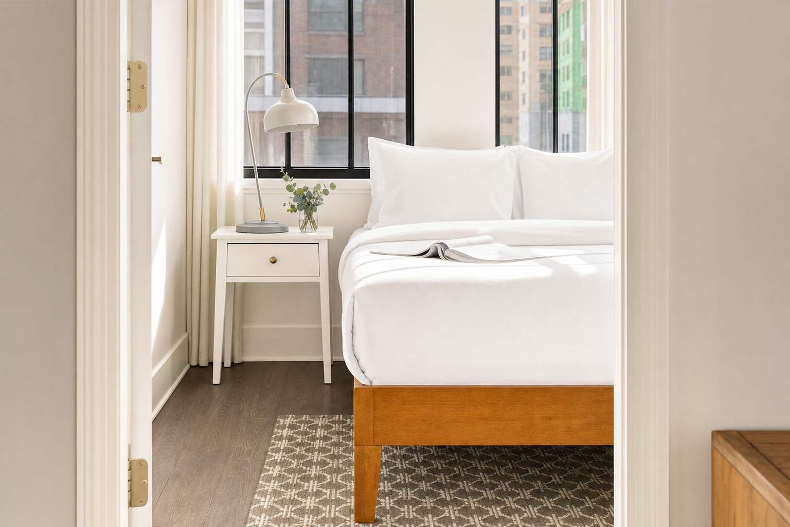A bedroom with a bed, a nightstand, a lamp, and a window with a view of buildings.