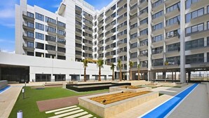 A modern multi-story building with a courtyard featuring a pool, palm trees, and a blue pathway. - Bloomfields Sleek 1BR in Azizi Mirage 1 (Dubai)