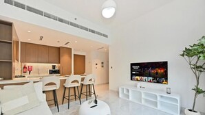A modern living room with a sofa, a TV, a kitchen area, and a dining space. - Bloomfields Sleek 1BR in Azizi Mirage 1 (Dubai)