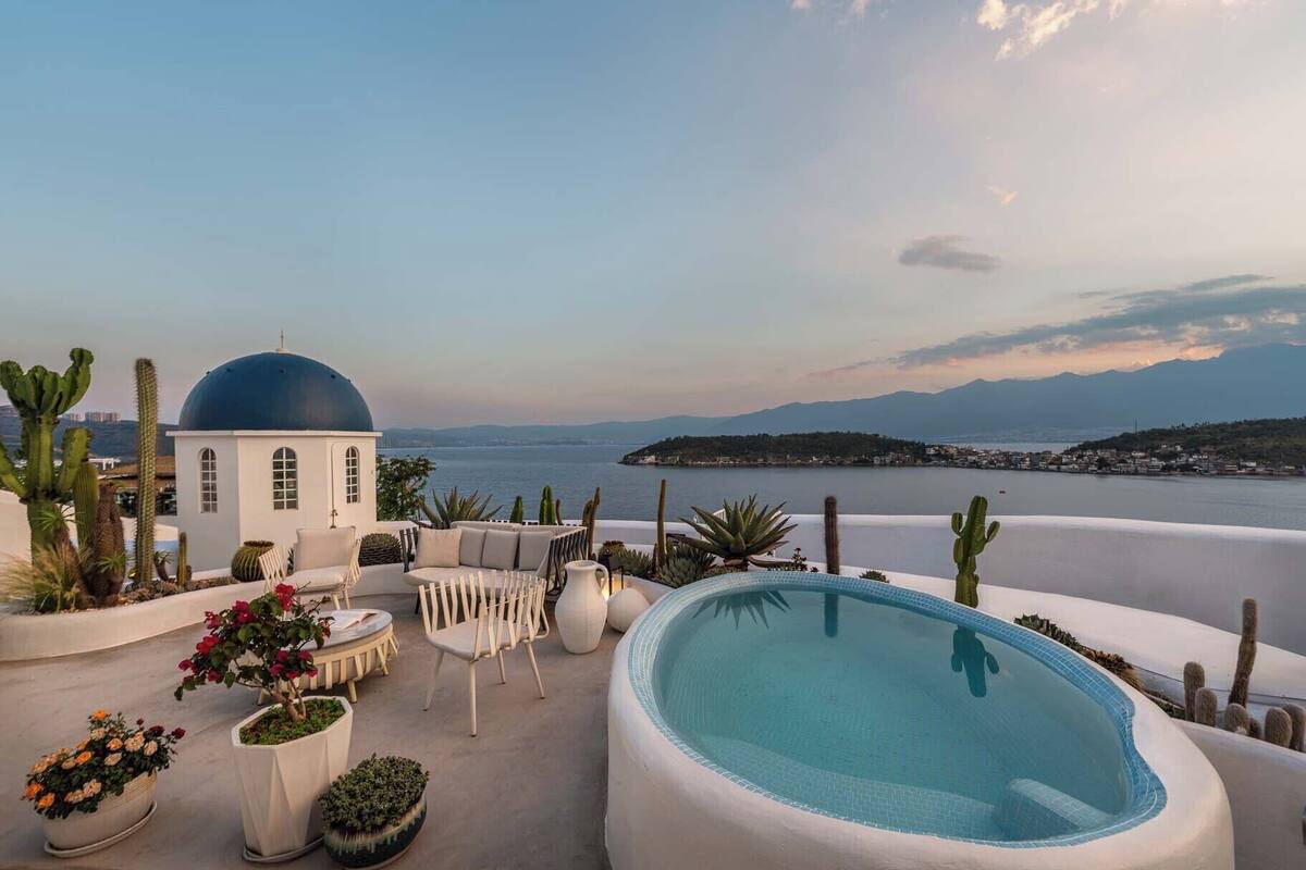 Une terrasse sur le toit avec une piscine, des meubles blancs, des cactus et une vue sur la mer et les montagnes.