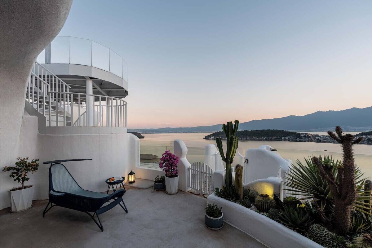 Un balcon donnant sur un plan d’eau et des montagnes, avec une balustrade blanche, une chaise et des plantes en pot.