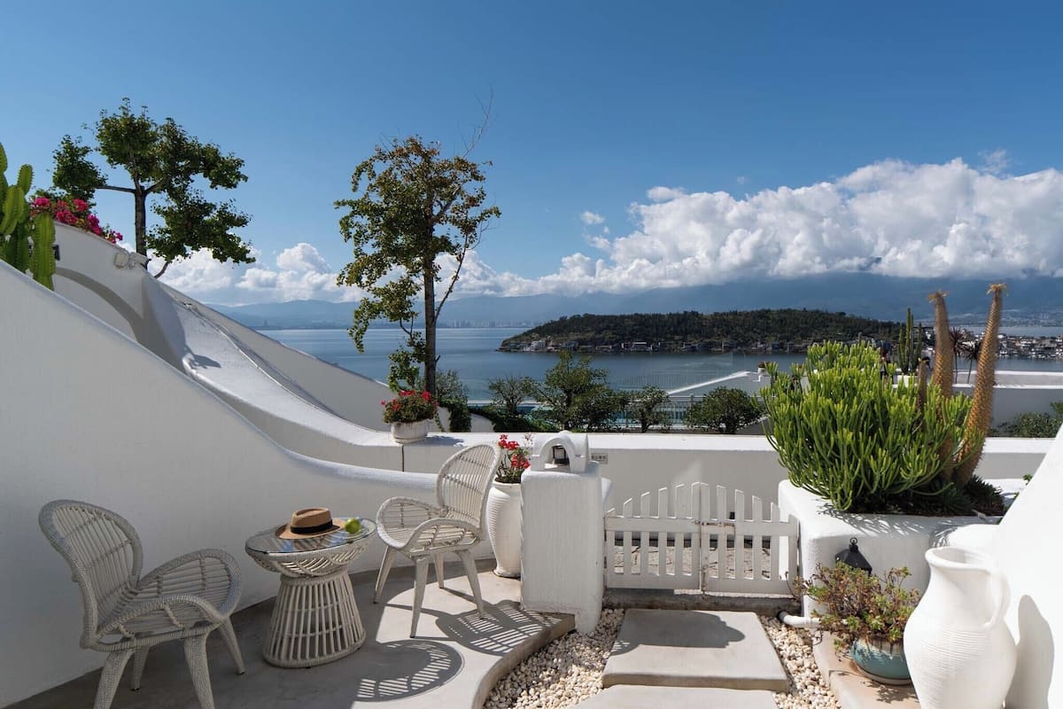 Une terrasse dotée de meubles en osier, d’une clôture blanche et offrant une vue sur la mer et le paysage lointain.