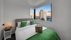 A hotel room with a bed, a nightstand, a lamp, a green blanket, a wicker basket, and a view of a cityscape through a large window. - Inviting Brisbane Family 2BR | CBD | Pool & Gym (Brisbane City)