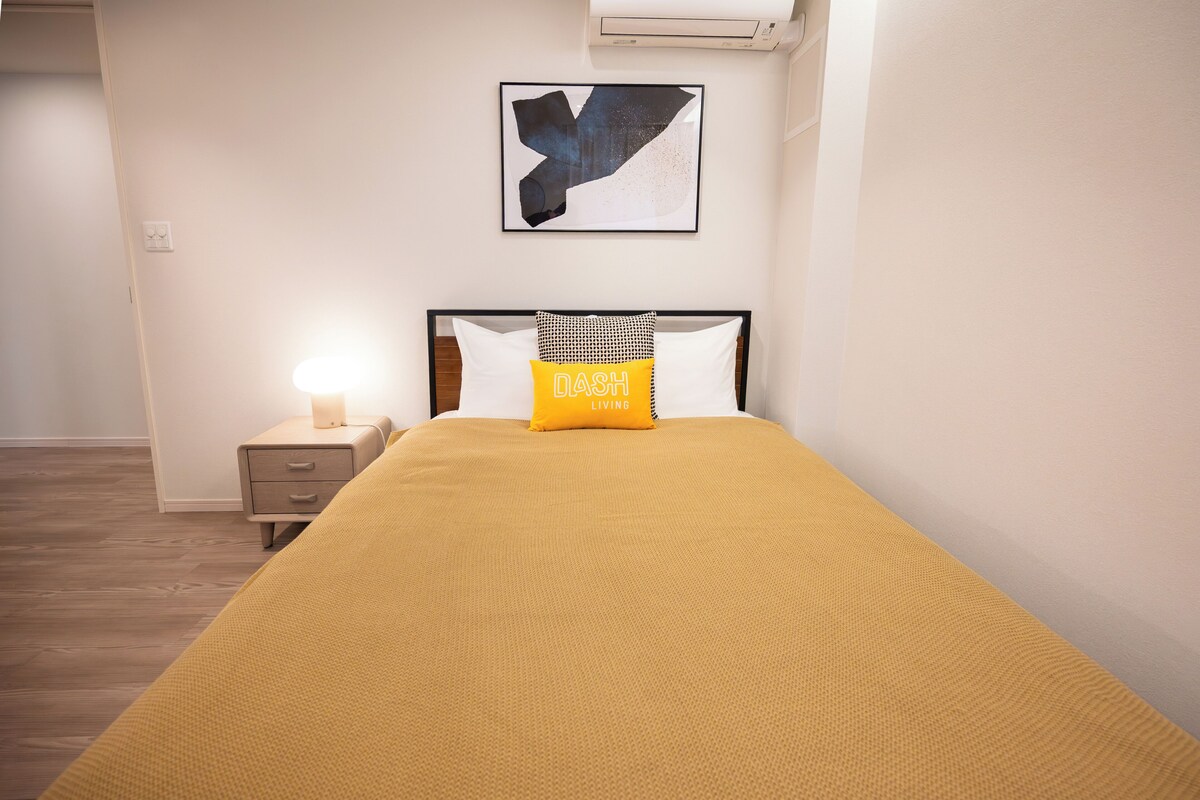 A neatly made bed with a yellow blanket, a black and white pillow, and a framed abstract artwork above it.