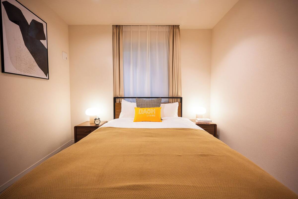 A hotel room with a bed, a nightstand with a lamp, a wall clock, and a window with curtains.