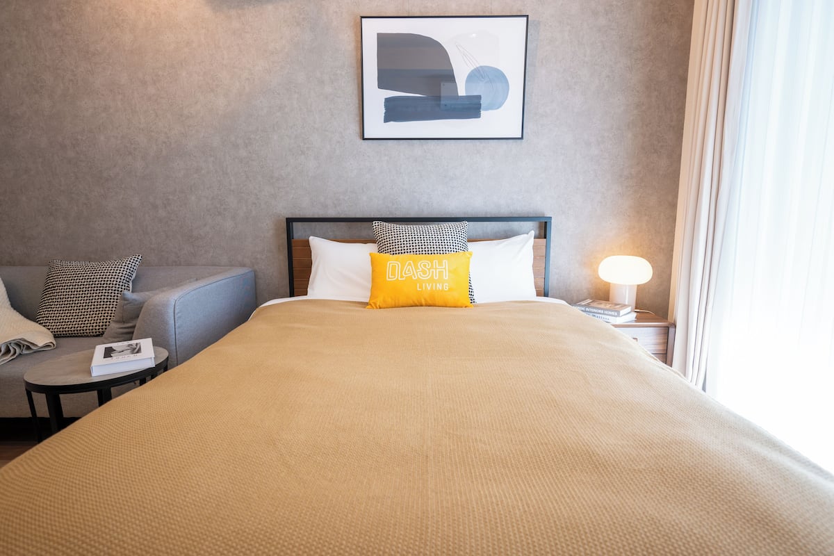 A hotel room with a bed, a sofa, a bedside table, a lamp, and a framed picture on the wall.
