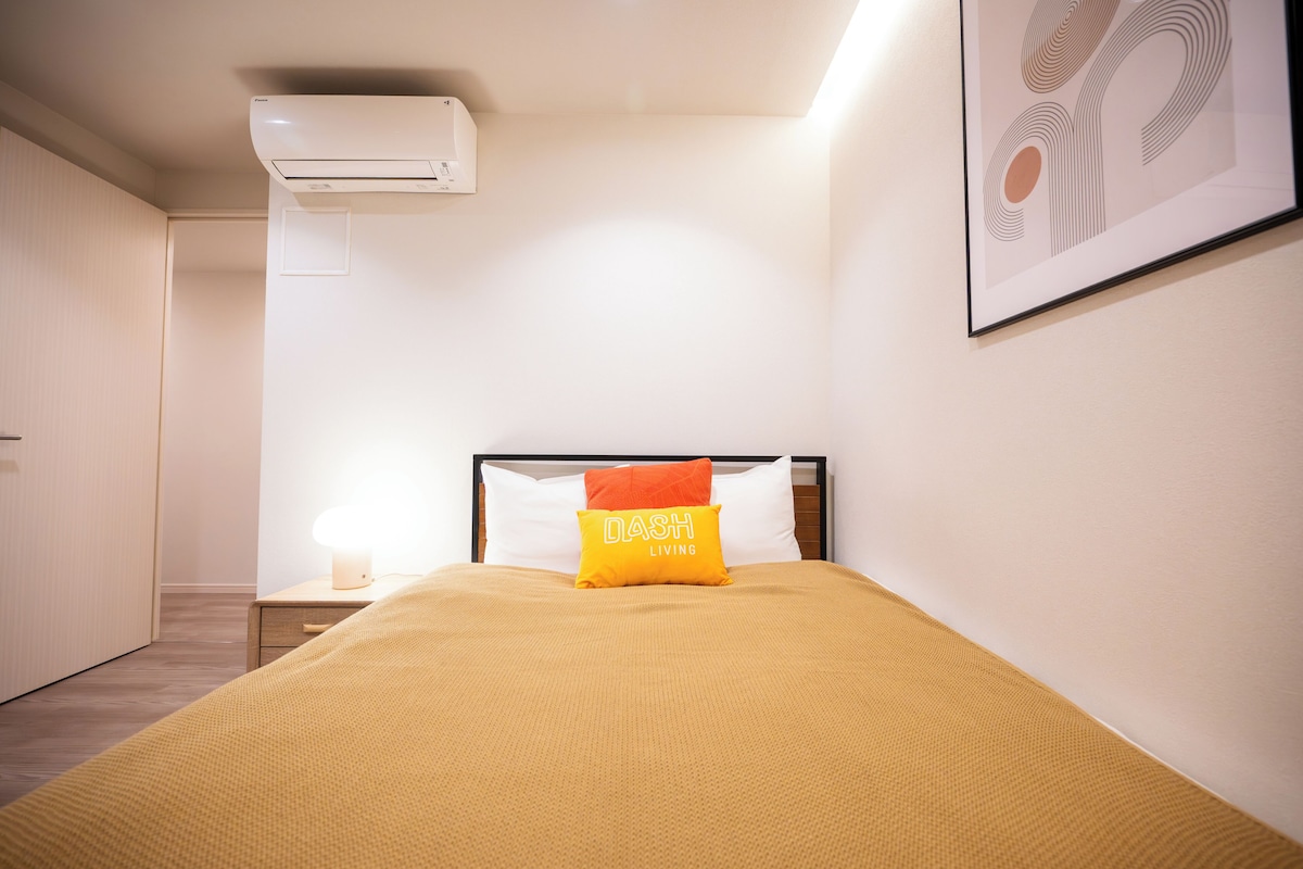A neatly made bed with a yellow blanket, a bedside lamp, and a pillow with 'DASH LIVING' text.