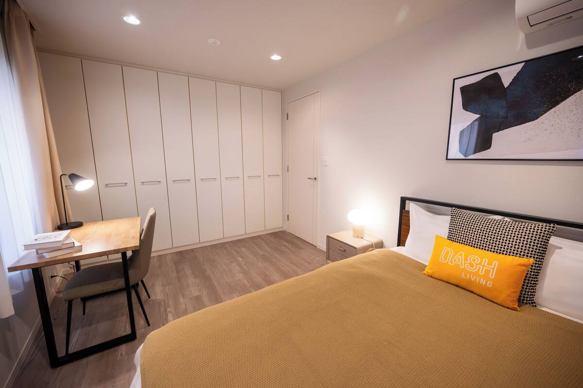 A bedroom with a bed, a desk, a chair, a nightstand, and a wall-mounted air conditioner.