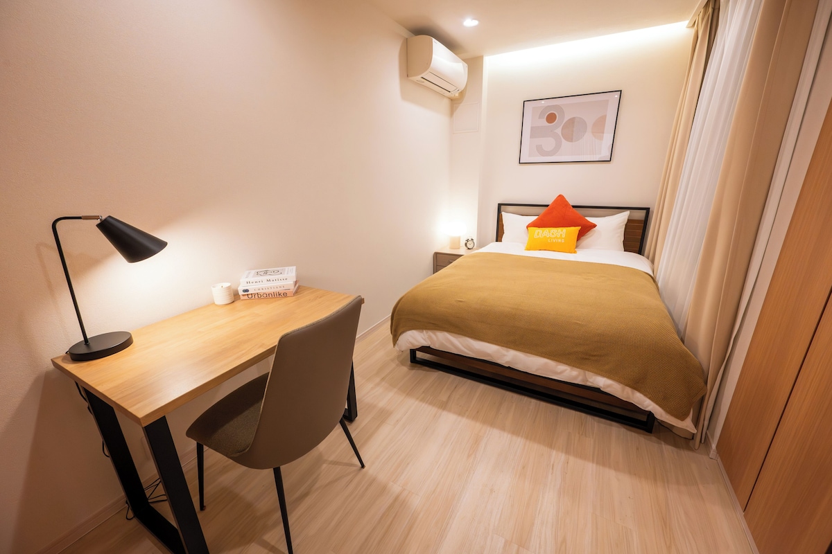 A hotel room with a bed, a desk with a chair, a lamp, an air conditioner, and a framed picture on the wall.