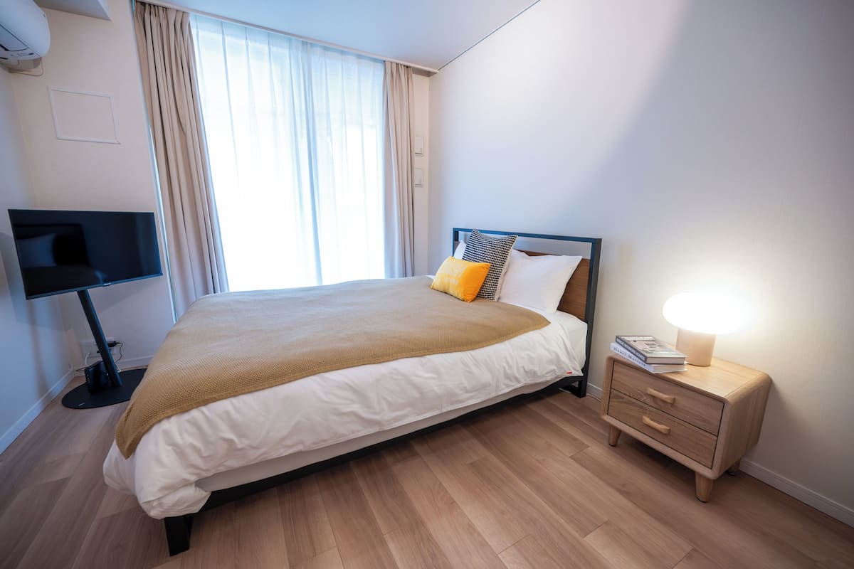 A neatly made bed with a brown blanket, a bedside table with a lamp, and a TV stand.