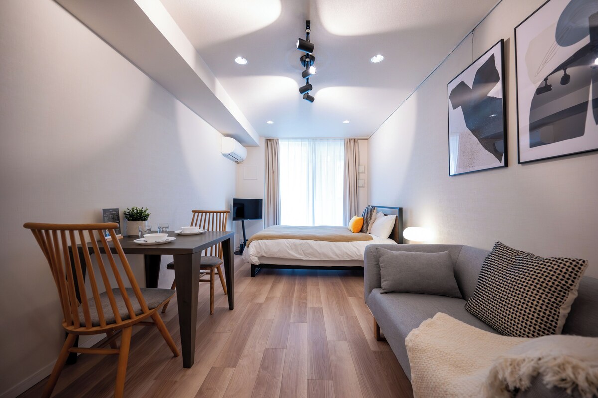A modern hotel room with a bed, a sofa, a dining table with chairs, and a TV.