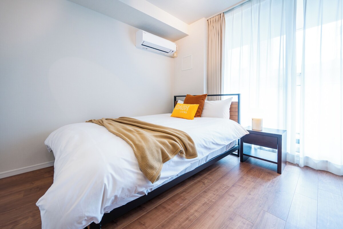 A bedroom with a bed, a bedside table, a lamp, an air conditioner, and a window with curtains.