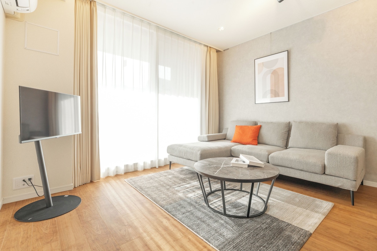 A modern living room with a flat-screen TV, a gray sofa with an orange pillow, a round coffee table, and a framed picture on the wall.