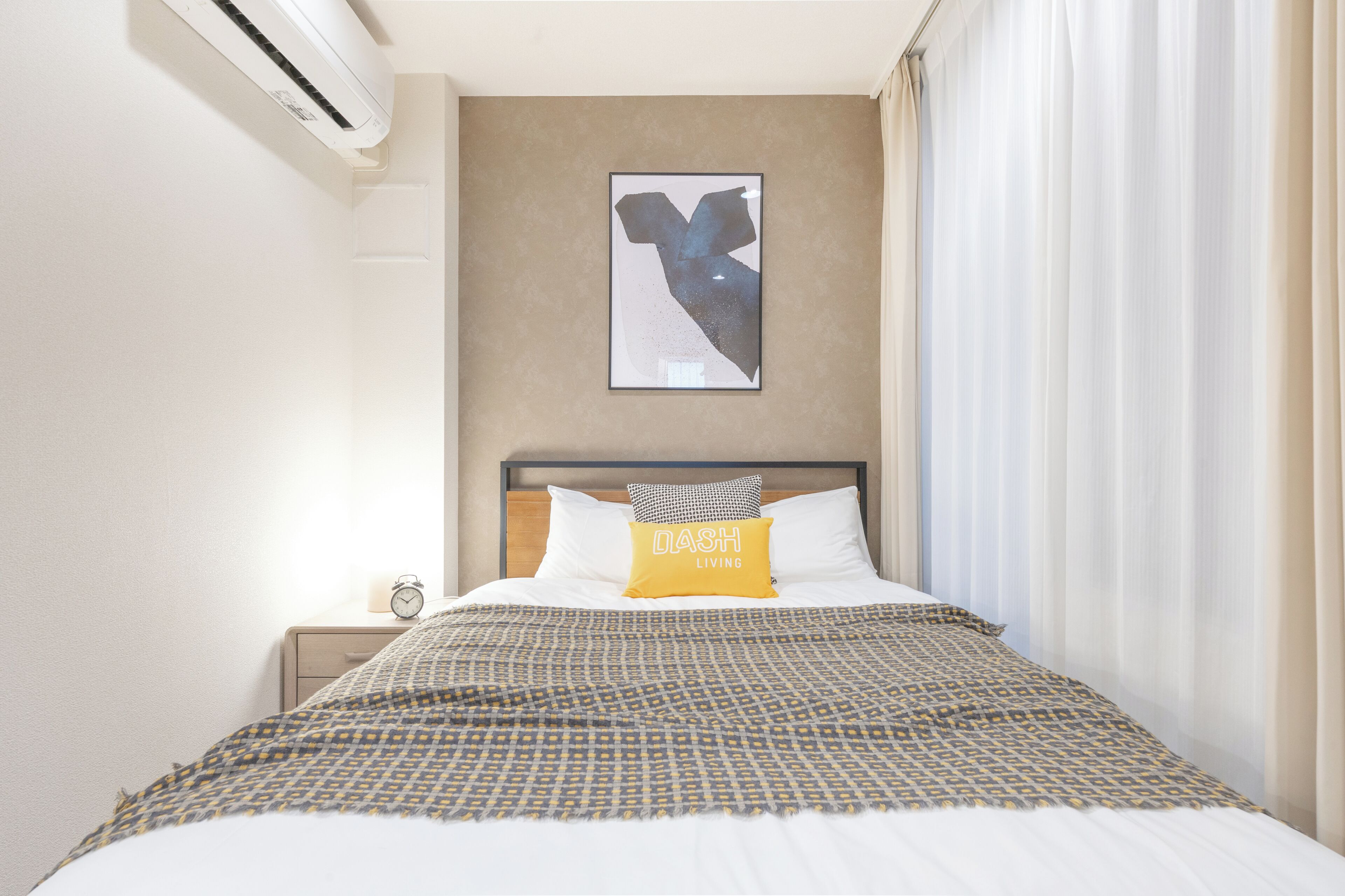 Una cama bien tendida con ropa de cama estampada, un aire acondicionado de pared, una obra de arte en un marco y una mesita de noche con un reloj.