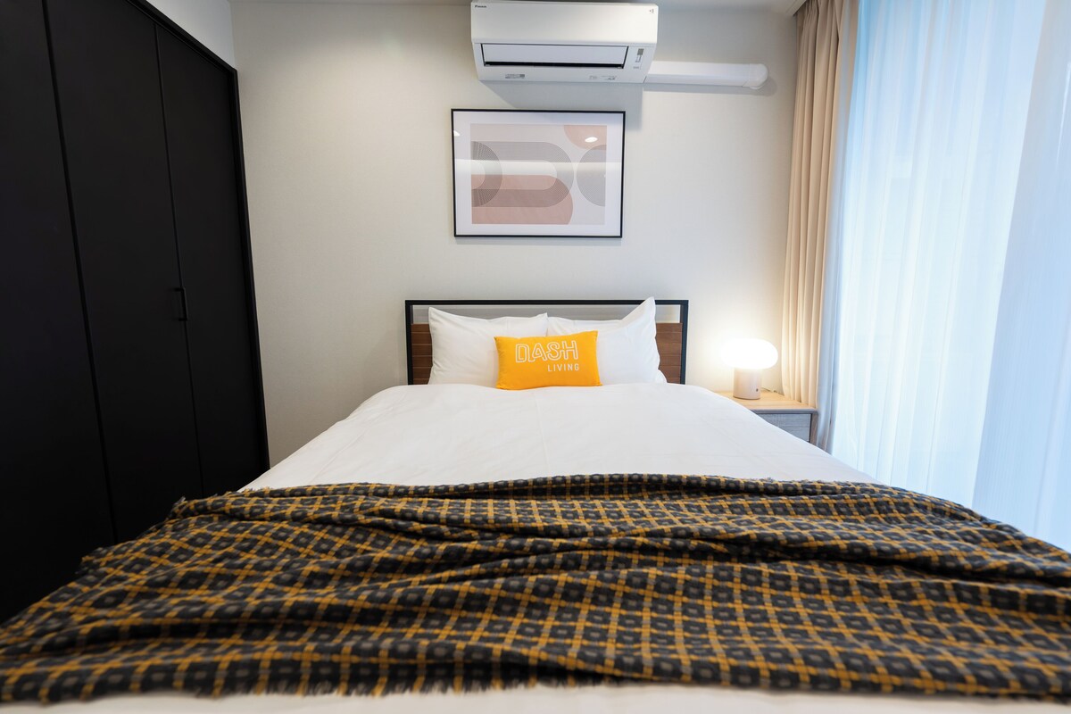 A hotel room with a bed, a nightstand, an air conditioning unit, a framed picture on the wall, and a window with curtains.