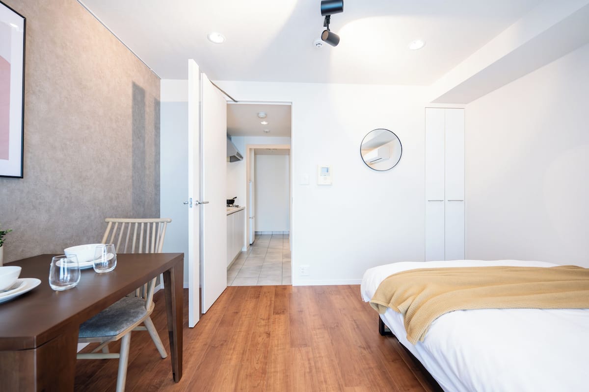 A modern hotel room with a wooden dining table, a bed with a beige blanket, a round mirror, and a wardrobe.