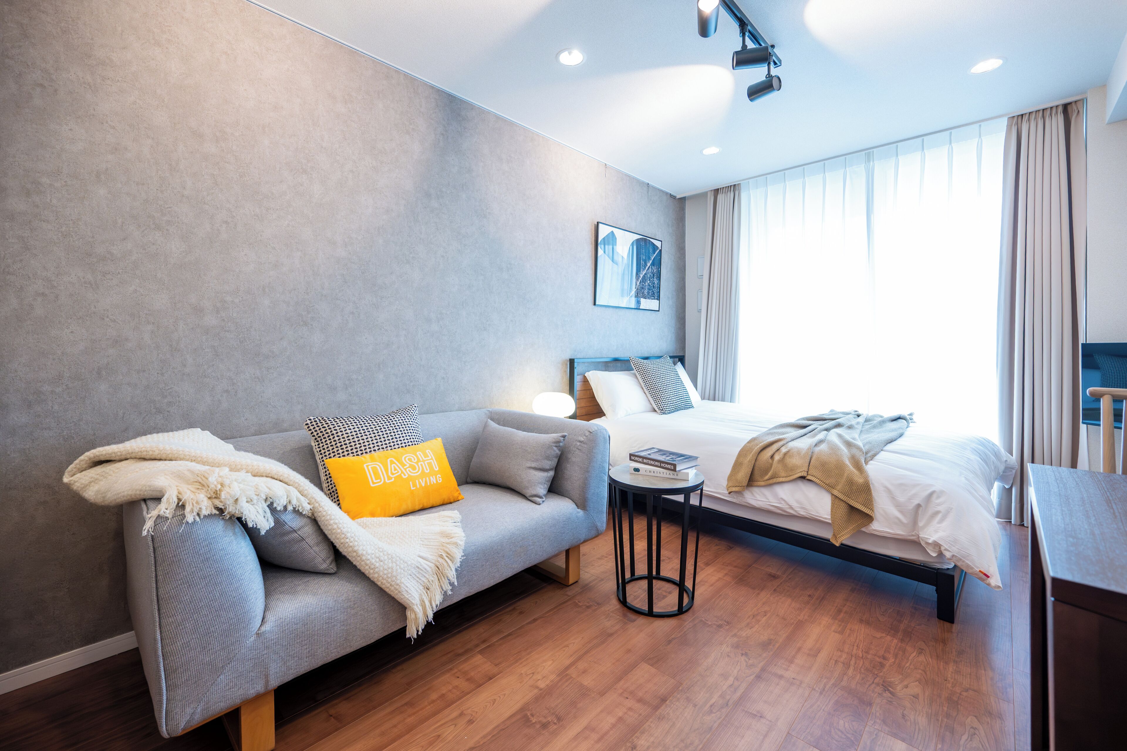 A modern hotel room with a grey sofa, a bed with white linens, a small round table, and a large window with curtains.
