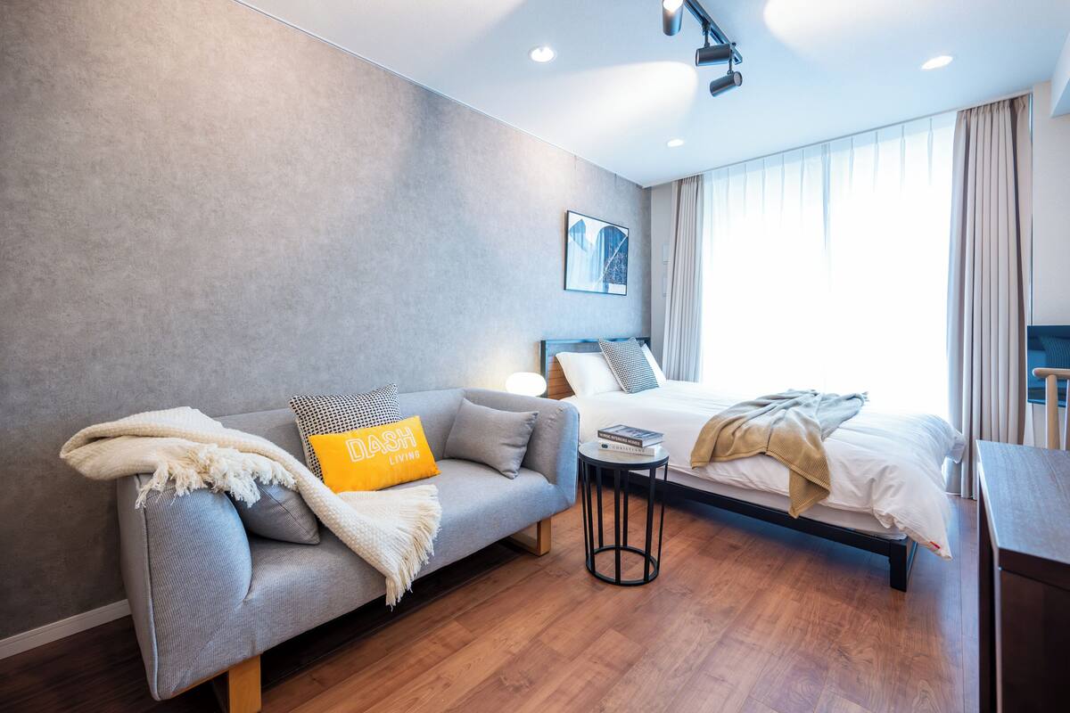 A modern hotel room with a grey sofa, a bed with white linens, a small round table, and a large window with curtains.