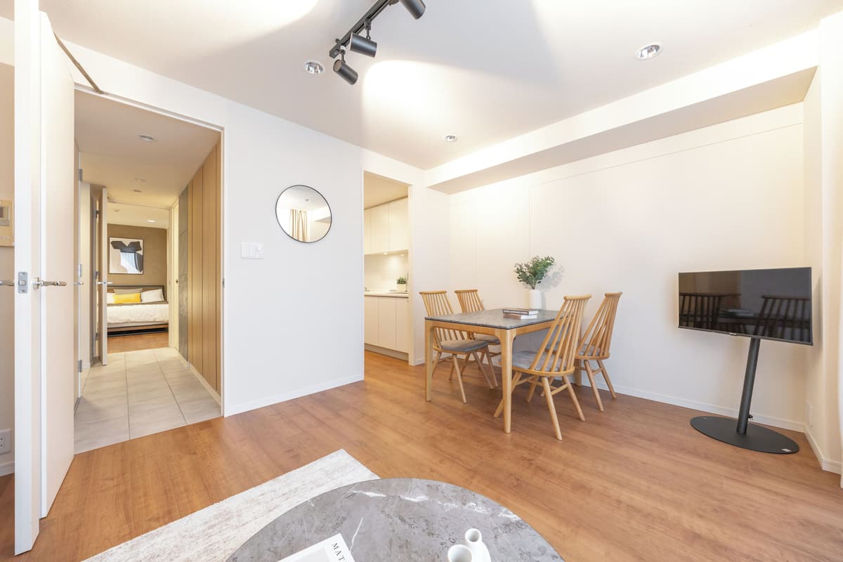 A modern living room with a dining area, a flat-screen TV, and a hallway leading to a bedroom.