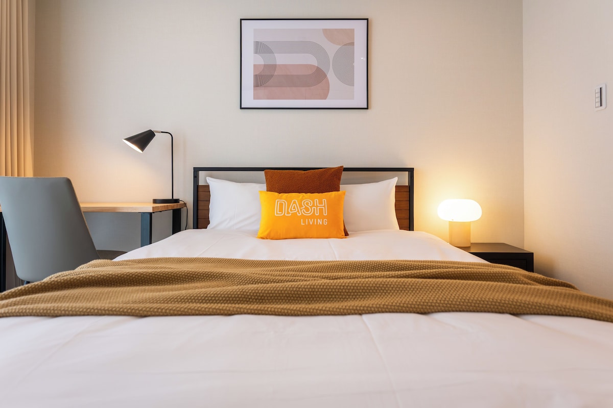 A neatly made bed with a brown blanket, a pillow with 'LASH LIVING' text, a lamp, and a framed abstract artwork on the wall.