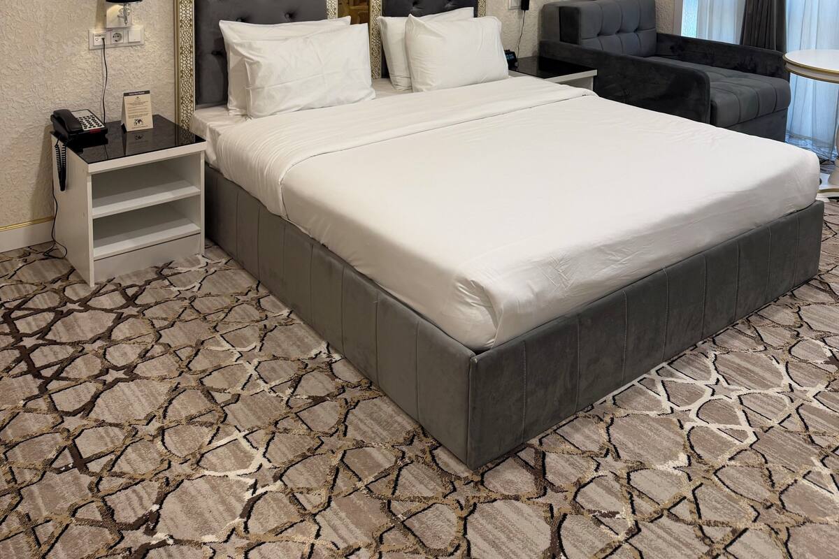 Un'ampia camera d'hotel con un letto grande, una poltrona e una scrivania. Sono presenti due lampade a parete e un telefono su un comodino. La stanza ha un tappeto decorato e un soffitto dal design originale.