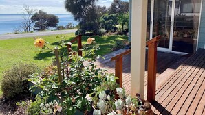 A wooden deck with a glass door leading to a covered porch, surrounded by a garden with various plants and a view of the sea. - Absolute waterfront views, a cosy, country style retreat, set amongst the trees. (Newhaven)