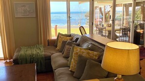 A living room with a sofa, a table, and a lamp. There is a window with a view of the ocean. - Absolute waterfront views, a cosy, country style retreat, set amongst the trees. (Newhaven)